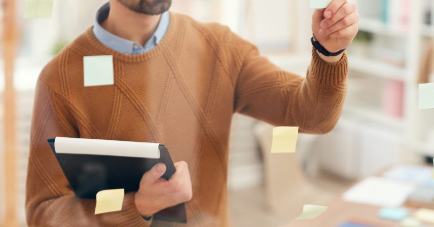 Mid-section portrait of bearded man placing sticker notes on glass wall while planning project in office, copy space
