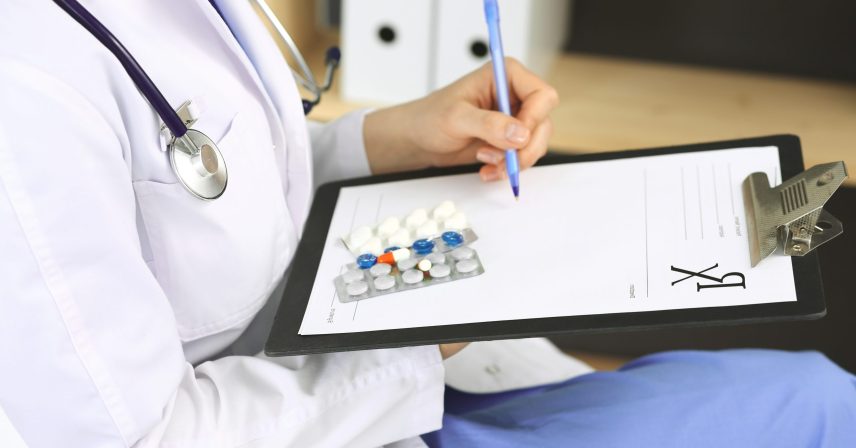 Unknown woman doctor at work at hospital. Pills at medical clipboard. Young female physician write prescription or filling up medical form while sitting in hospital office, close-up.